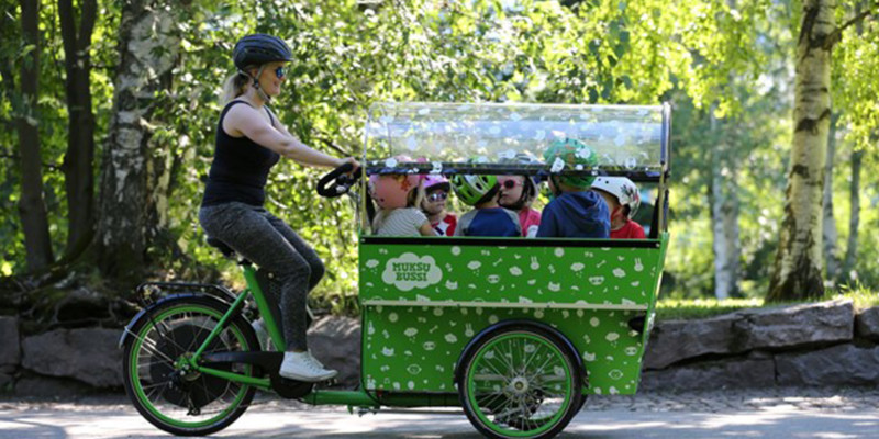 Little children sit in a cargo bike box, and a grown-up is riding the bike.