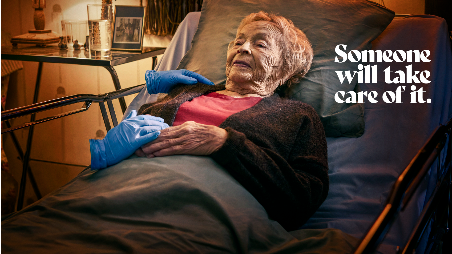 An elderly person lies on a hospital bed. A nurse's glove-covered hands touch the person softly. The nurse is invisible except for blue working gloves.