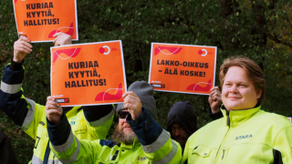 Huomiovaatteisiin pukeutuneita ammattiliitto JHL:n jäseniä lakkokyltit kädessään, syyskesäinen puumaisema taustalla.