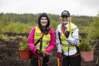 Kaksi tyttöä seisoo metsässä taimiämpäreiden kanssa ja katsoo kameraan nauraen