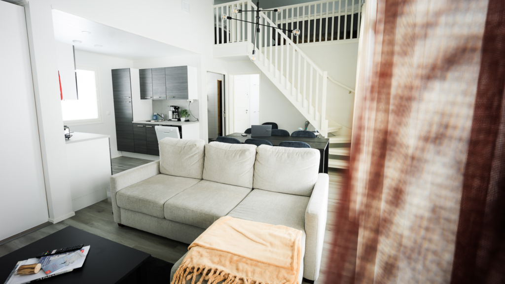 A view of the living room and kitchen in one of JHL's holiday houses. There is a grey sofa in the foreground, and in the background there is a large dining table and a spacious kitchen with black cabinets.