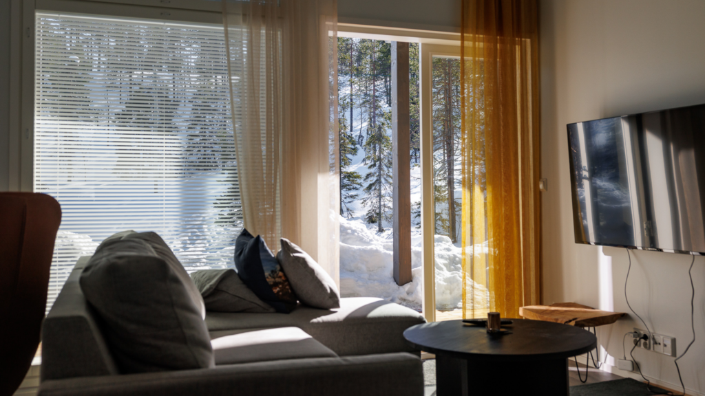 A photo taken from the living room of one of JHL's holiday houses in Ruka. There is a grey sofa and a small black coffee table in the foreground, and behind them is a window showing a bright, snowy forest scenery.