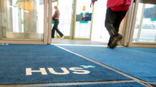 People walking out through automatic doors and a large blue doormat with text “HUS”.