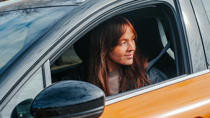 Tummatukkainen aikuinen istuu oranssissa autossa.