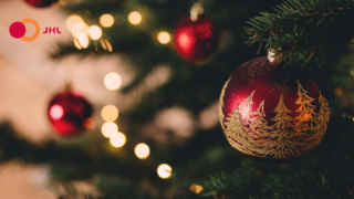 A close-up photo of a red and gold bauble in a Christmas tree and JHL's logo in the upper left corner.