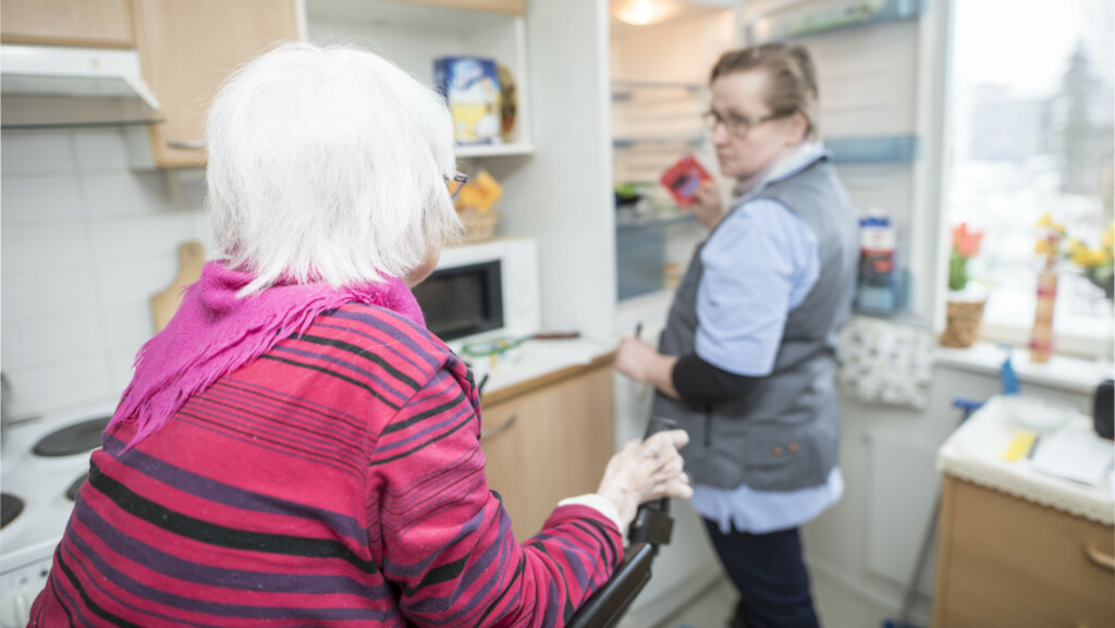 Valokuva vanhuksesta ja kotihoitajasta vanhuksen omassa keittiössä. Valkohiuksinen vanhus nojailee kuvan etualalla rollaattoriin selkä kameraan päin. Kuvassa taaempana hoitaja avaa jääkaappia.